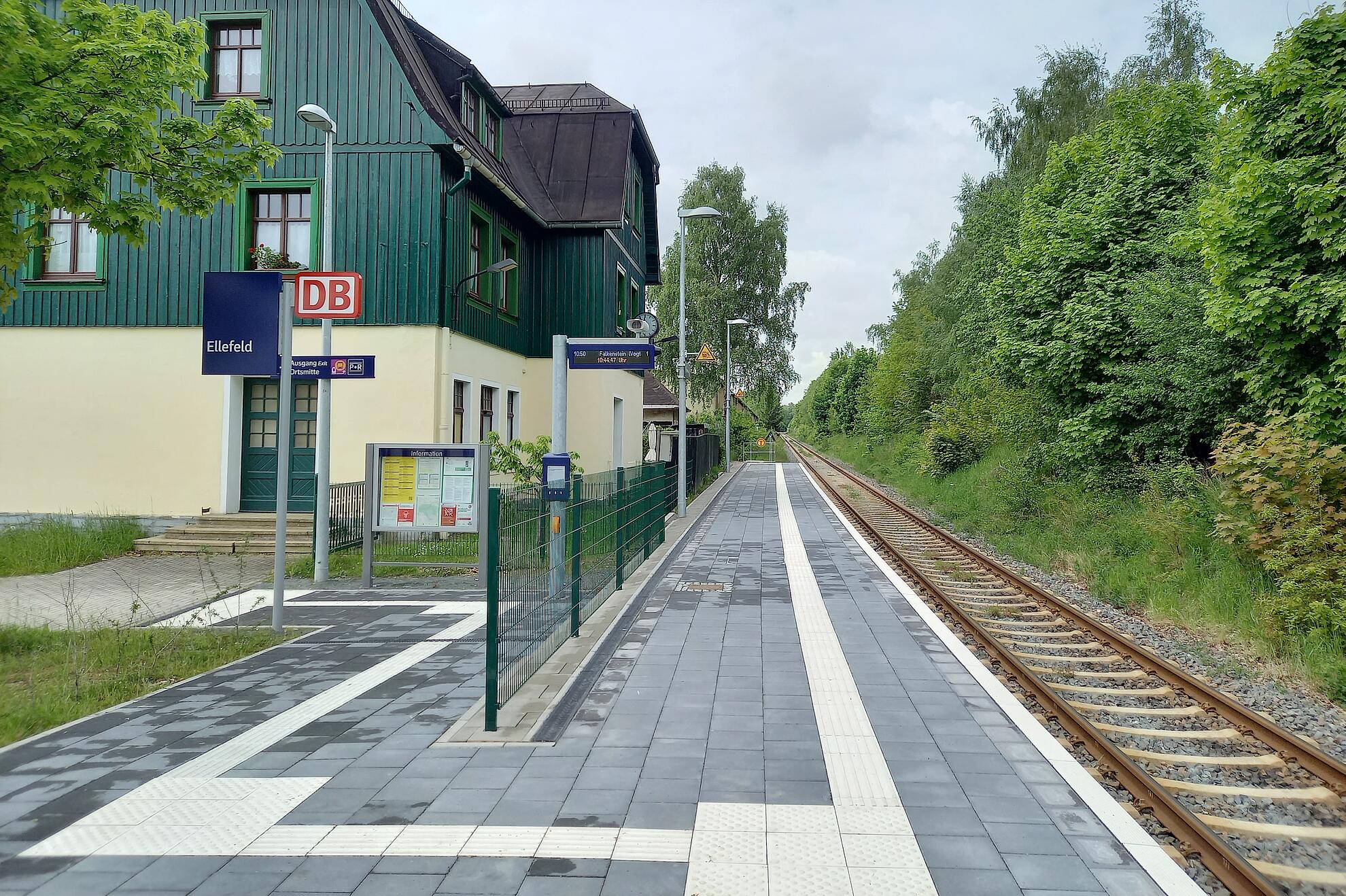 Bahnhof Ellefeld mit modernisiertem Bahnsteig, taktilen Leitstreifen und Gleis, umgeben von Bäumen und dem Stationsgebäude.