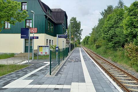 Bahnhof Ellefeld mit modernisiertem Bahnsteig, taktilen Leitstreifen und Gleis, umgeben von Bäumen und dem Stationsgebäude.