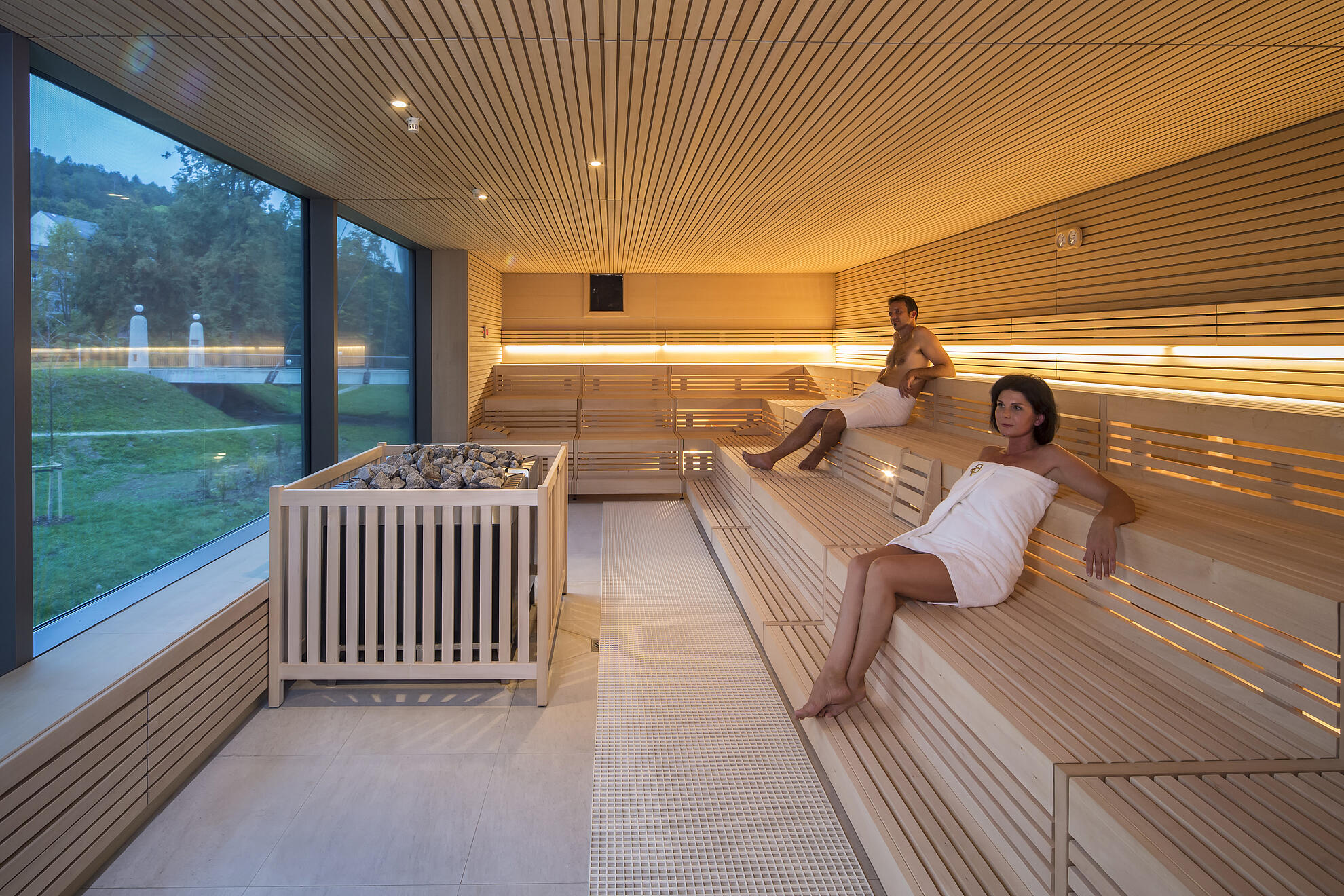 Moderne Panorama-Sauna mit hellen Holzbänken, warmen Lichtleisten und großem Fenster mit Blick ins Grüne. Eine Frau und ein Mann entspannen auf den Saunabänken, der Saunaofen mit Steinen steht im Vordergrund.