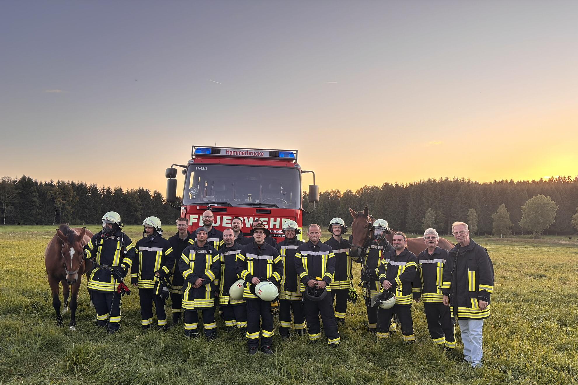 „Ein rotes Feuerwehrauto steht auf einem Feld. Davor Feuerwehrleute mit Pferd bei einer Übung.