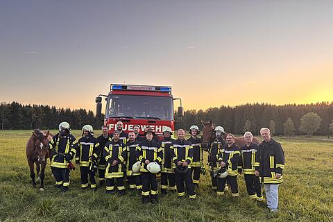 „Ein rotes Feuerwehrauto steht auf einem Feld. Davor Feuerwehrleute mit Pferd bei einer Übung.