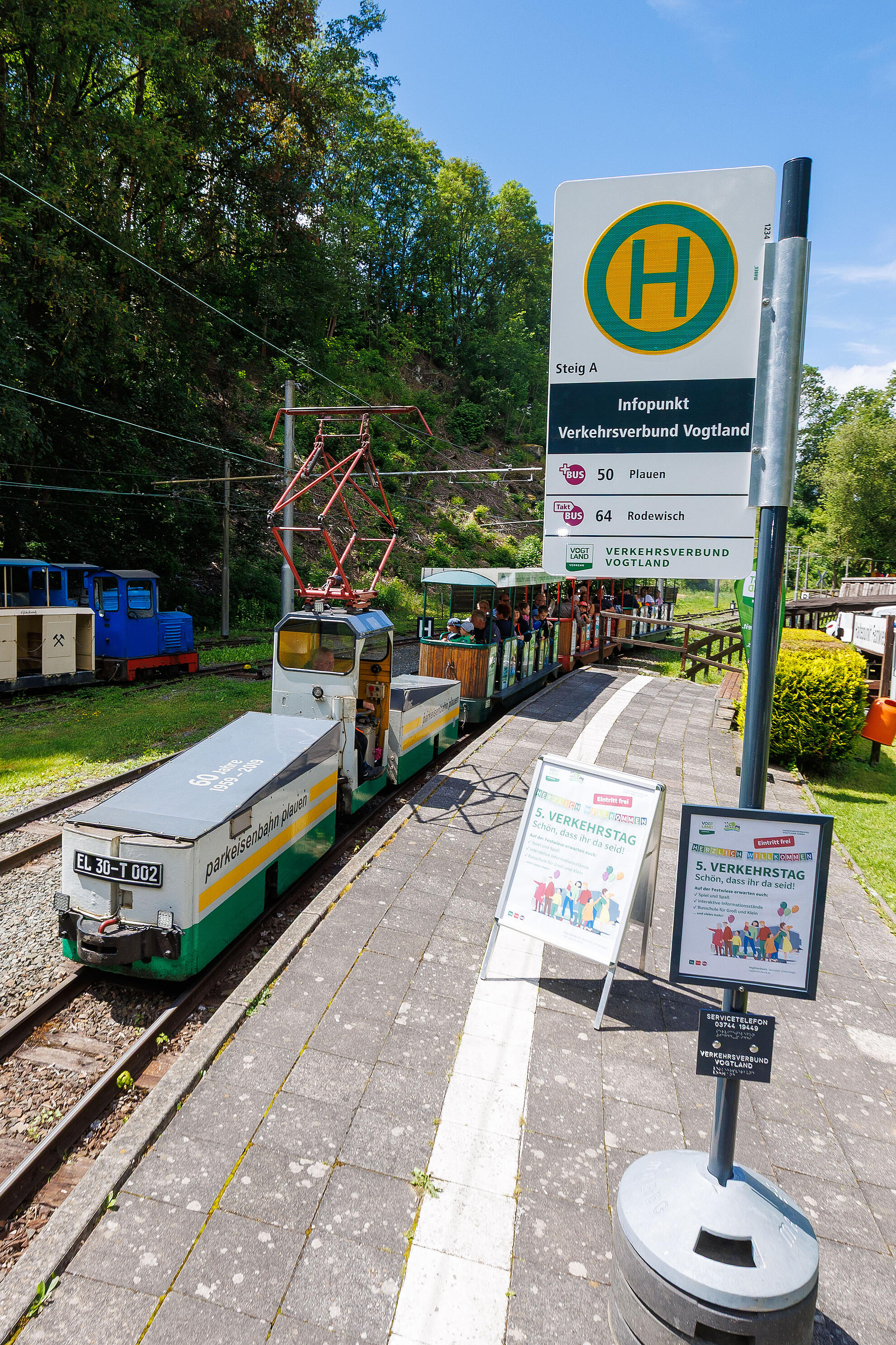 Kleine elektrische Parkeisenbahn mit offenen Wagen steht am Bahnsteig neben einem grünen Haltestellenschild, umgeben von Bäumen.