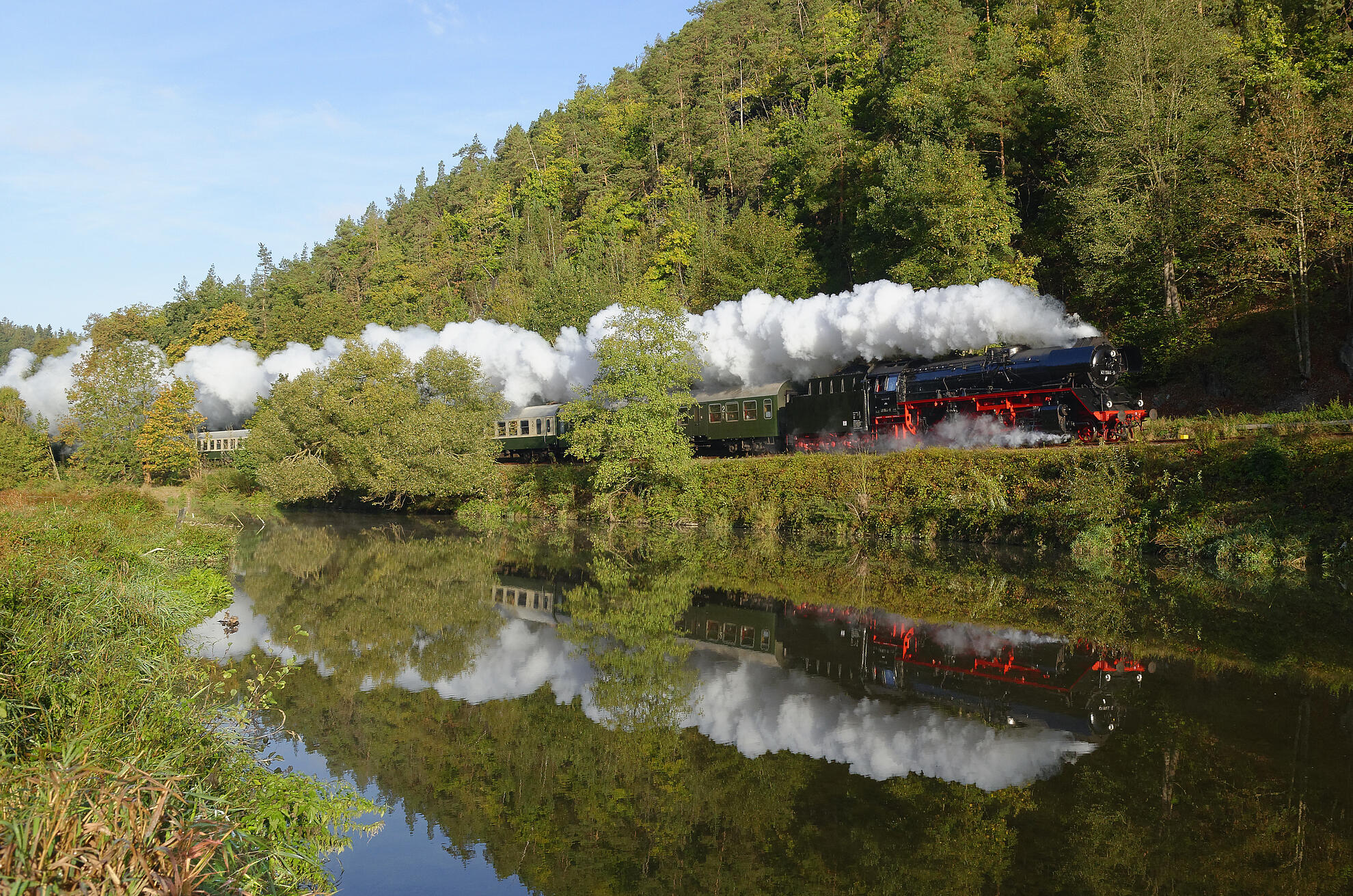 Ein Zug mit Dampflok fährt im Elstertal entlang des Flusses.