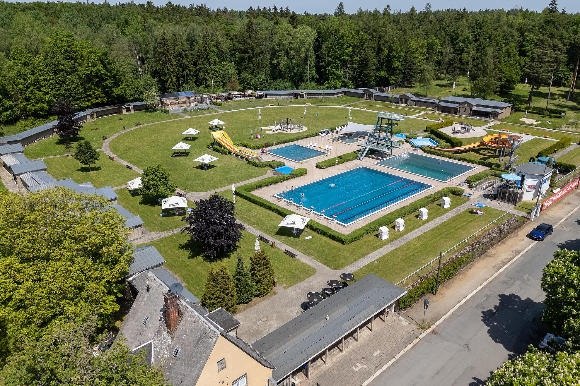 Ein großes Freibad mit mehreren Becken, Rutschen, Sprungturm und umliegenden Grünflächen aus der Vogelperspektive.