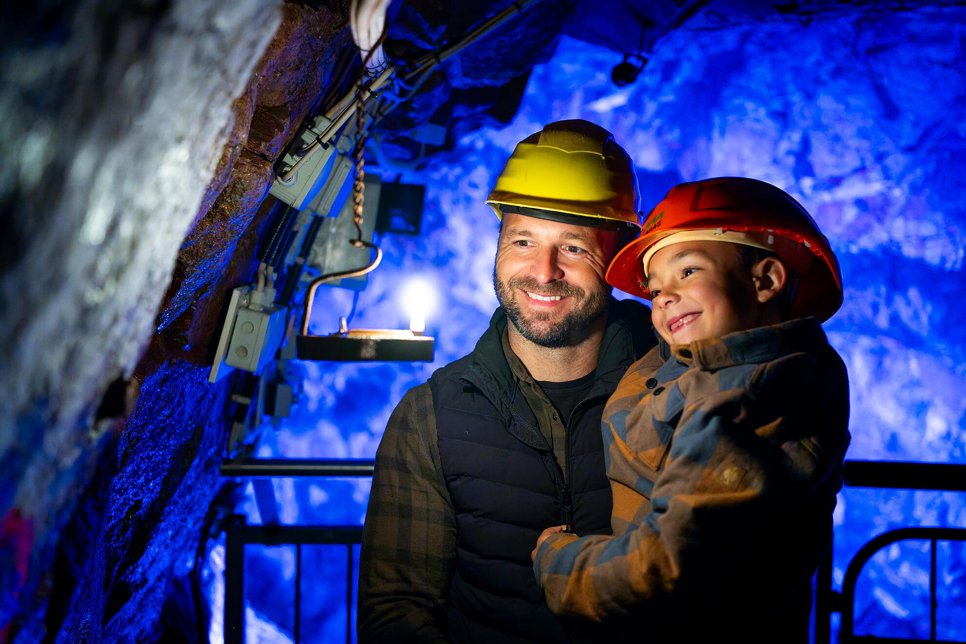 Ein Vater und sein Sohn mit Schutzhelmen lächeln bei einem Besuch unter Tage in der Grube Tannenberg. Das blaue Licht beleuchtet die Felswände des historischen Bergwerks.