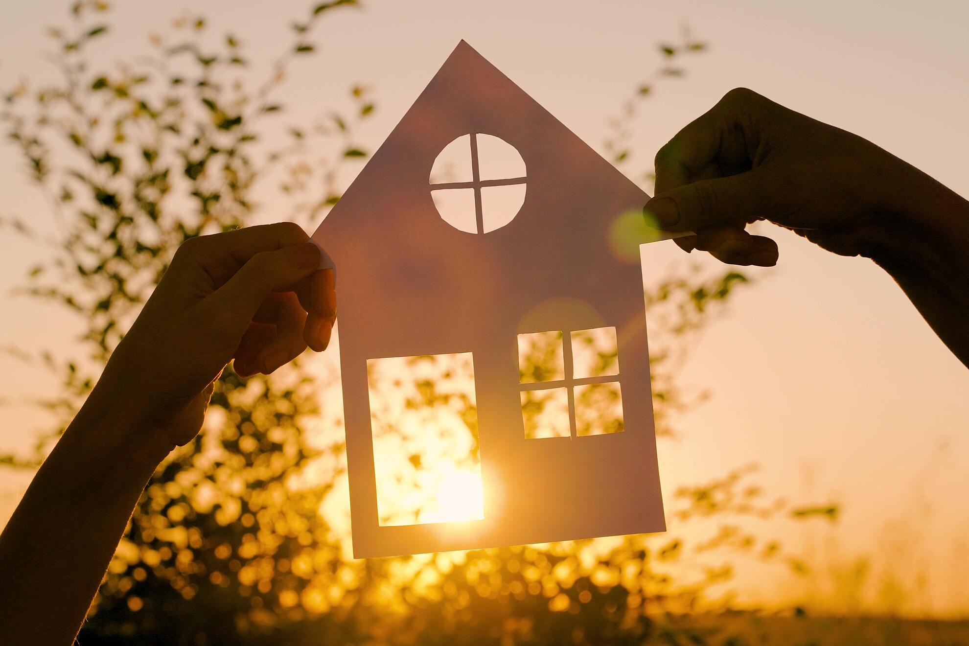 Zwei Hände halten ein kleines Haus aus Papier gegen das Licht, sodass die Konturen des ausgeschnittenen Hausmotivs hell durchscheinen.