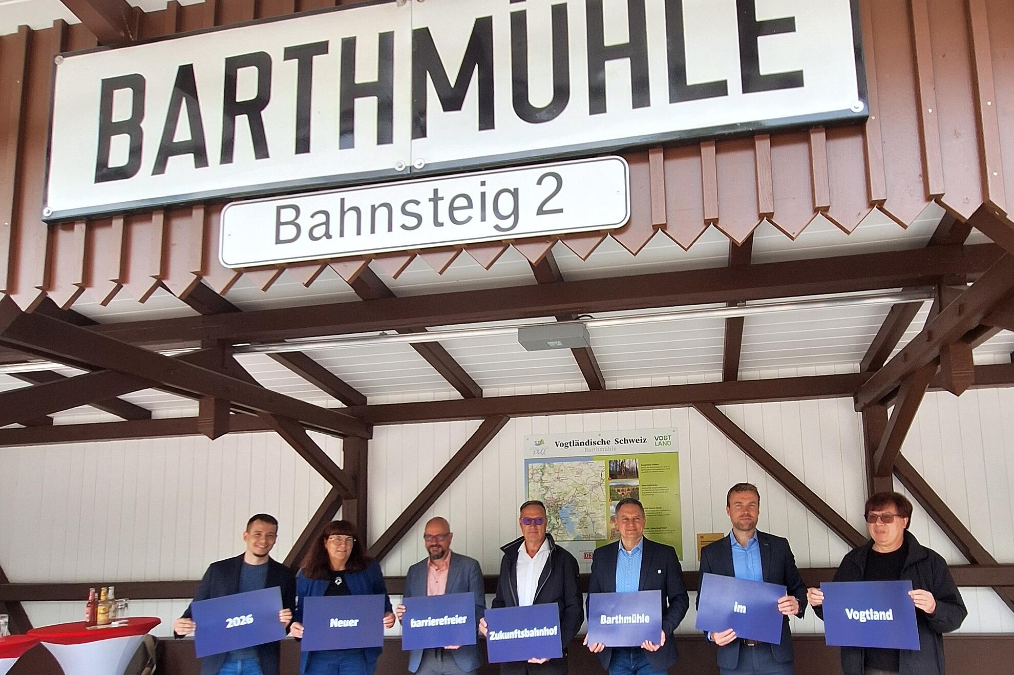 Sieben Personen stehen am Bahnhof Barthmühle und halten blaue Schilder zum neuen barrierefreien Zukunftsbahnhof 2026.