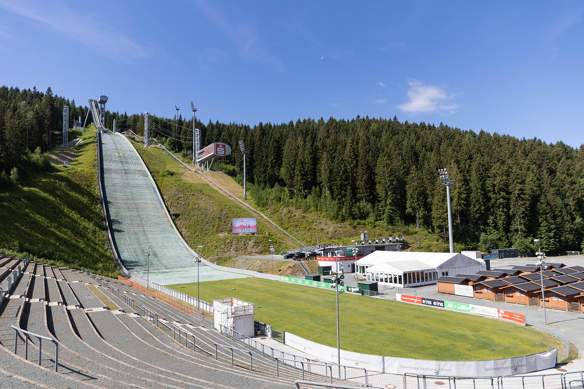 Sparkasse Vogtland Arena. Skisprungschanze in einer bewaldeten Hügellandschaft mit grüner Auslaufzone, Zuschauertribünen und Sportgebäuden unter blauem Himmel.