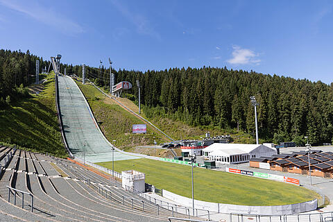 Sparkasse Vogtland Arena. Skisprungschanze in einer bewaldeten Hügellandschaft mit grüner Auslaufzone, Zuschauertribünen und Sportgebäuden unter blauem Himmel.
