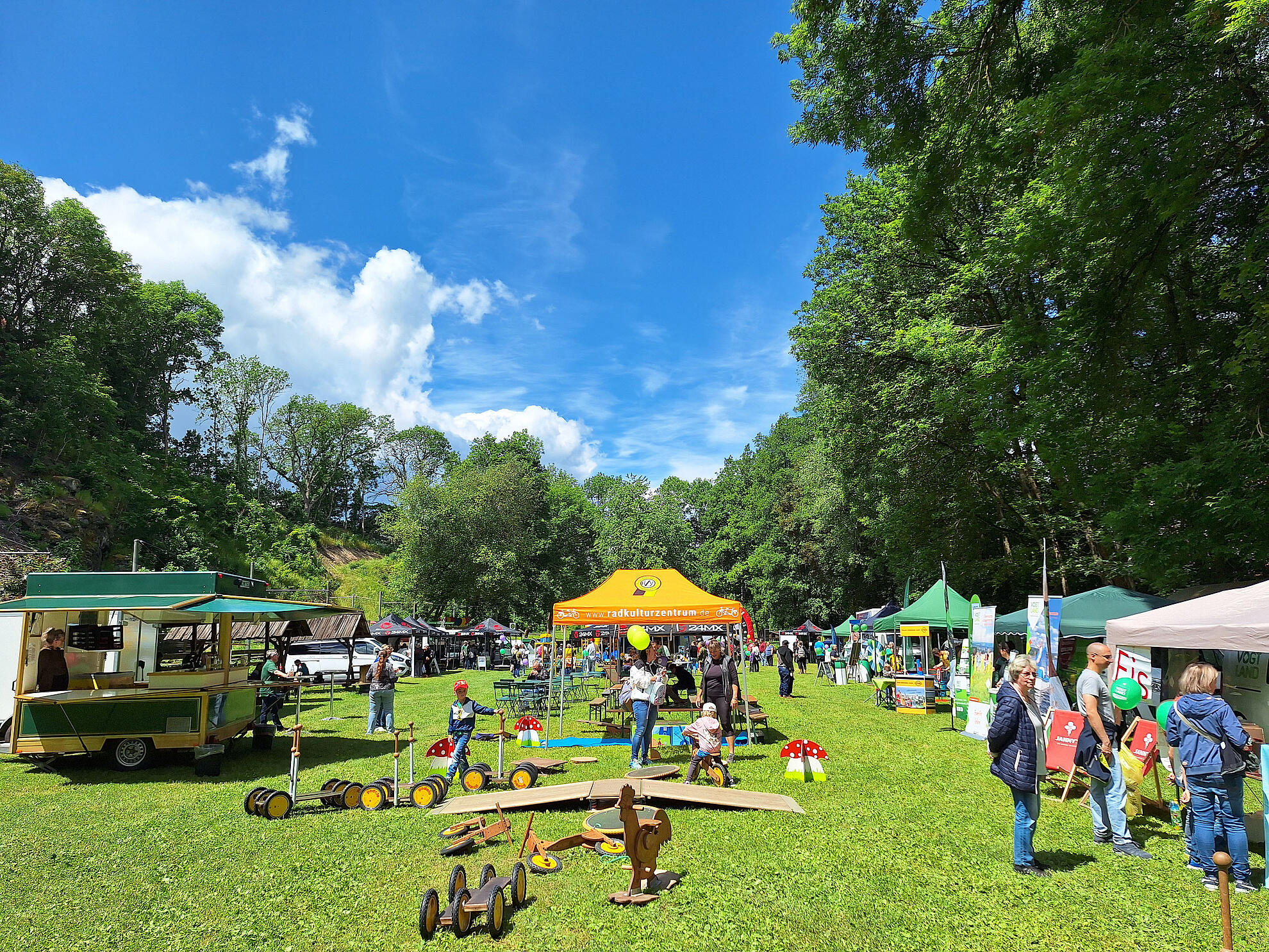 Outdoor‑Fest auf einer Wiese mit Ständen, Spielzeug aus Holz und vielen Besuchern vor blauem Himmel.