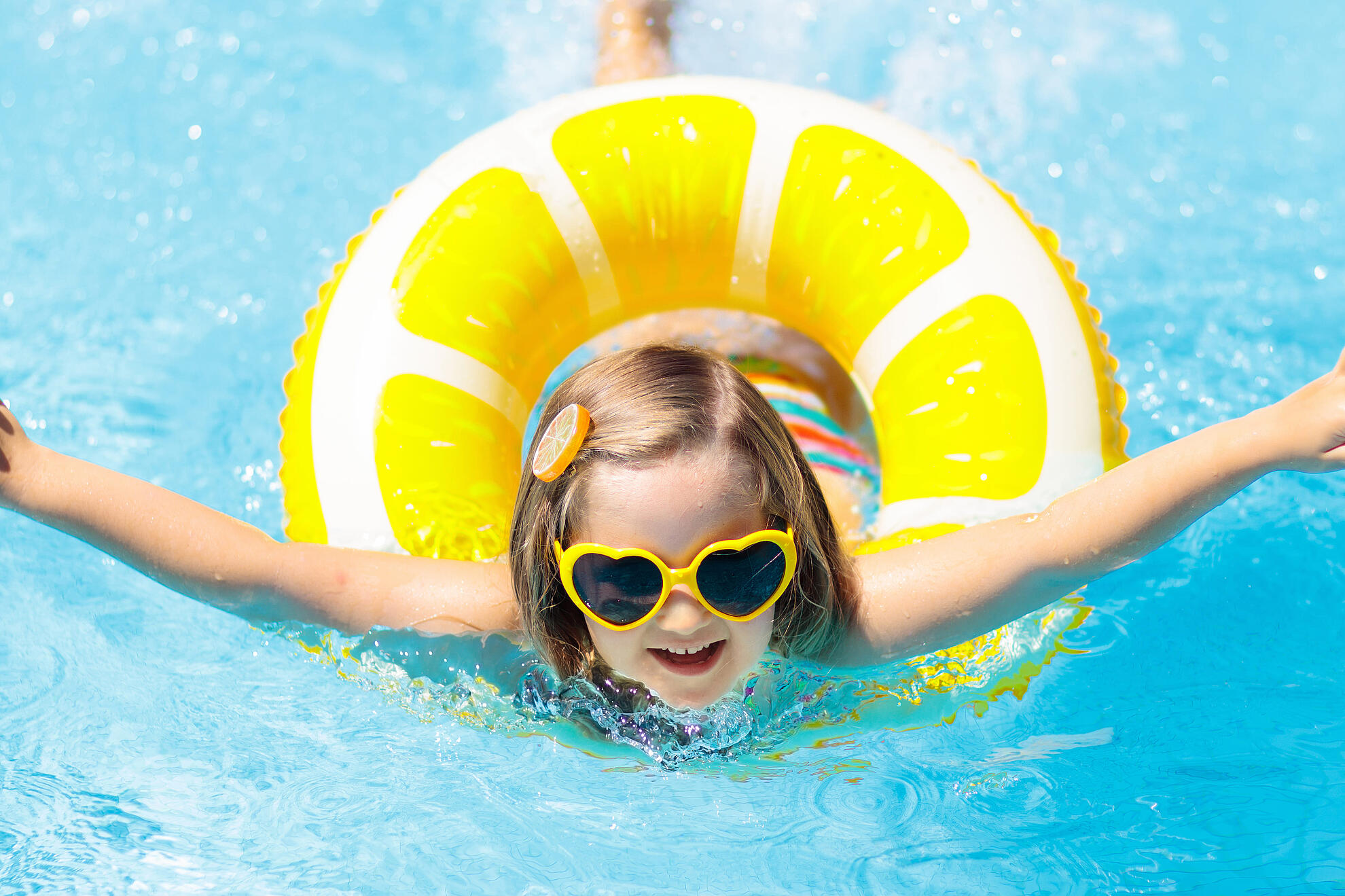 Ein Kind spielt im klaren blauen Pool, sitzt in einem gelben Zitronen‑Schwimmring und trägt gelbe herzförmige Sonnenbrille.
